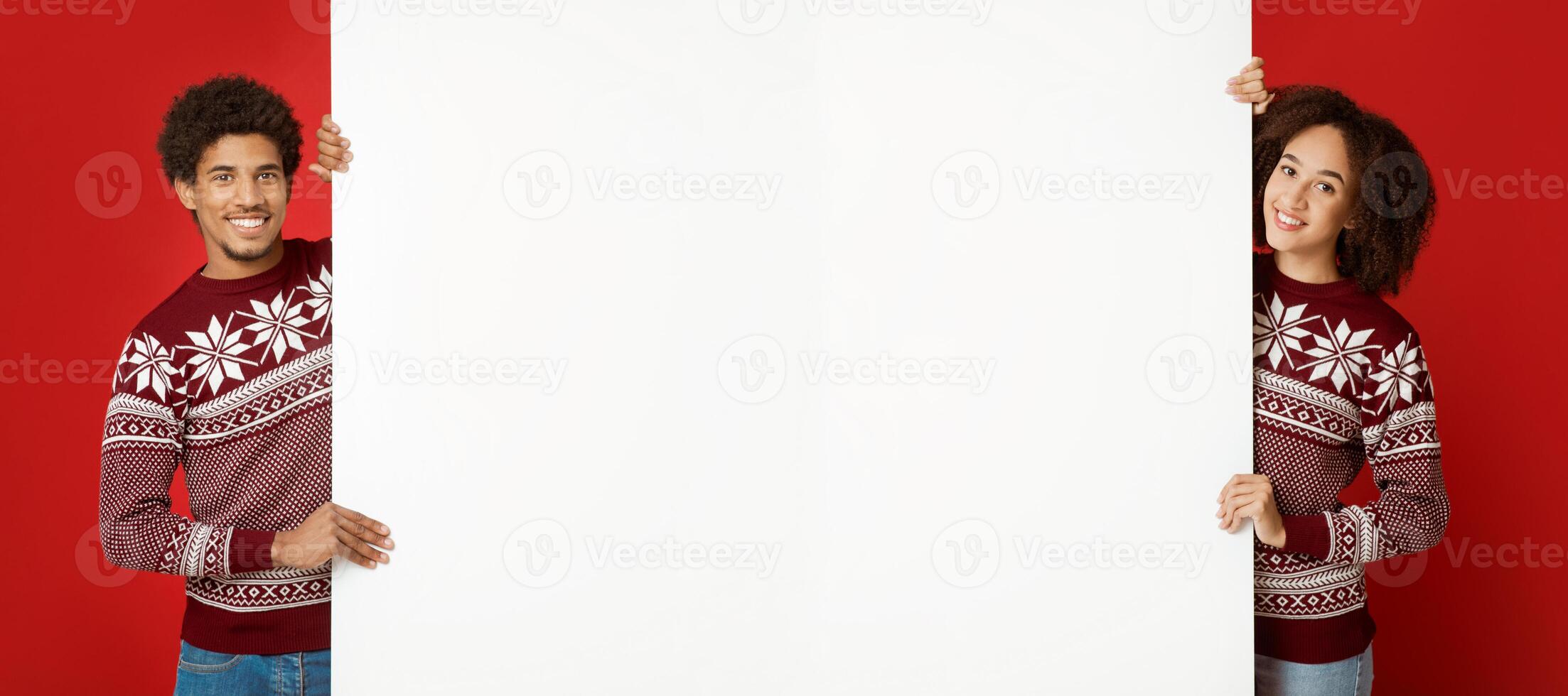 Smiling pretty young african american couple friends in same sweaters with pattern hold big white empty billboard and look at camera, mock up, copy space, isolated on red background, studio shot photo