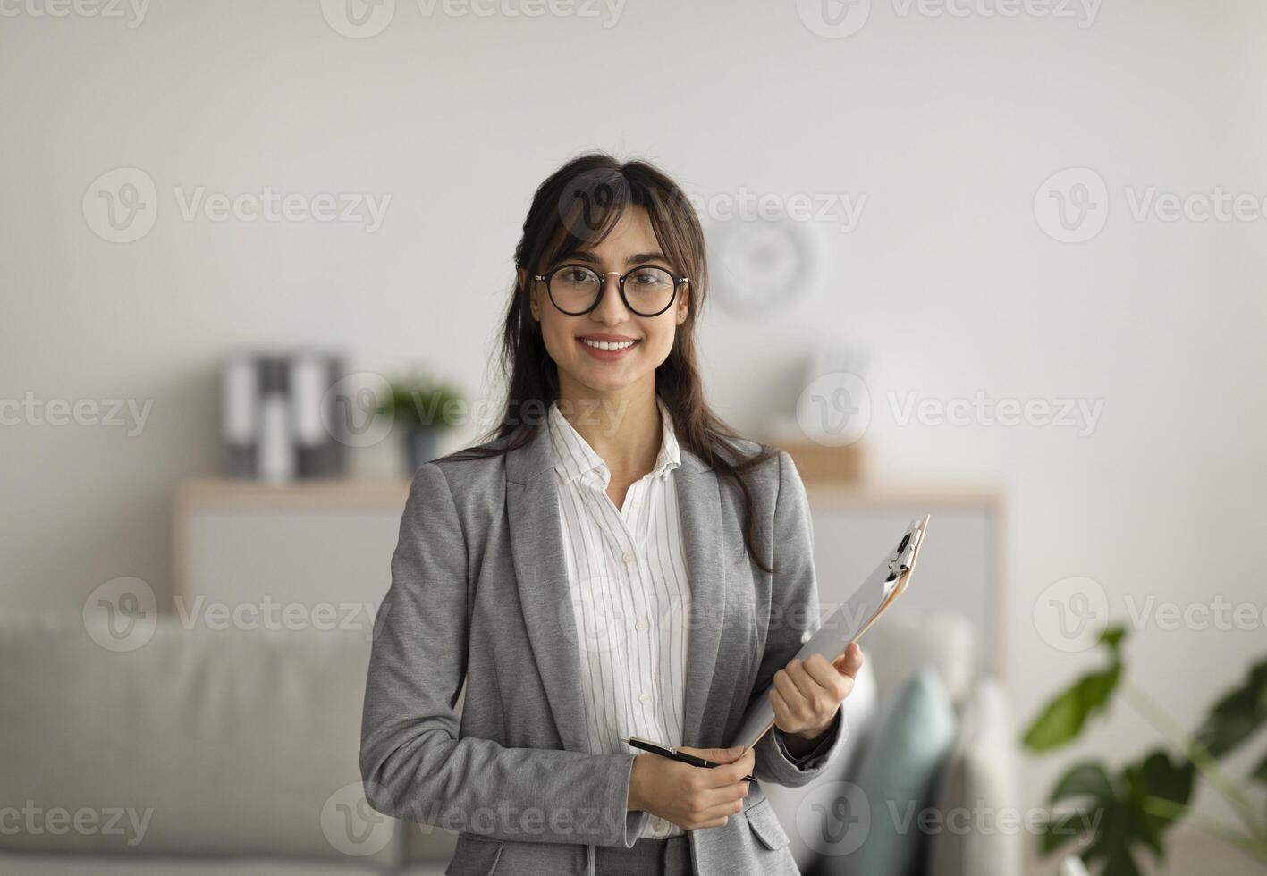 Portrait of cheerful female psychologist posing with clipboard, looking and smiling at camera, working in modern office. Psychotherapy services, mental health professional concept photo