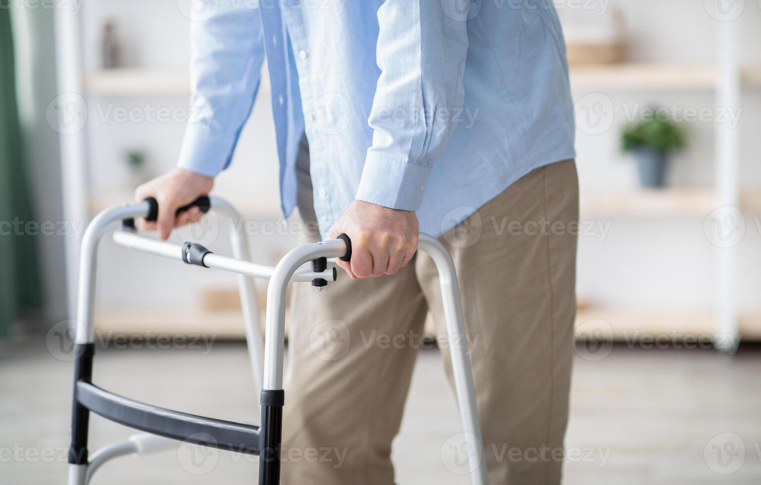 Cropped view of elderly man walking with frame at home, closeup. Unrecognizable senior male using medical equipment to move around his house. Disabled older person in need of professional help photo