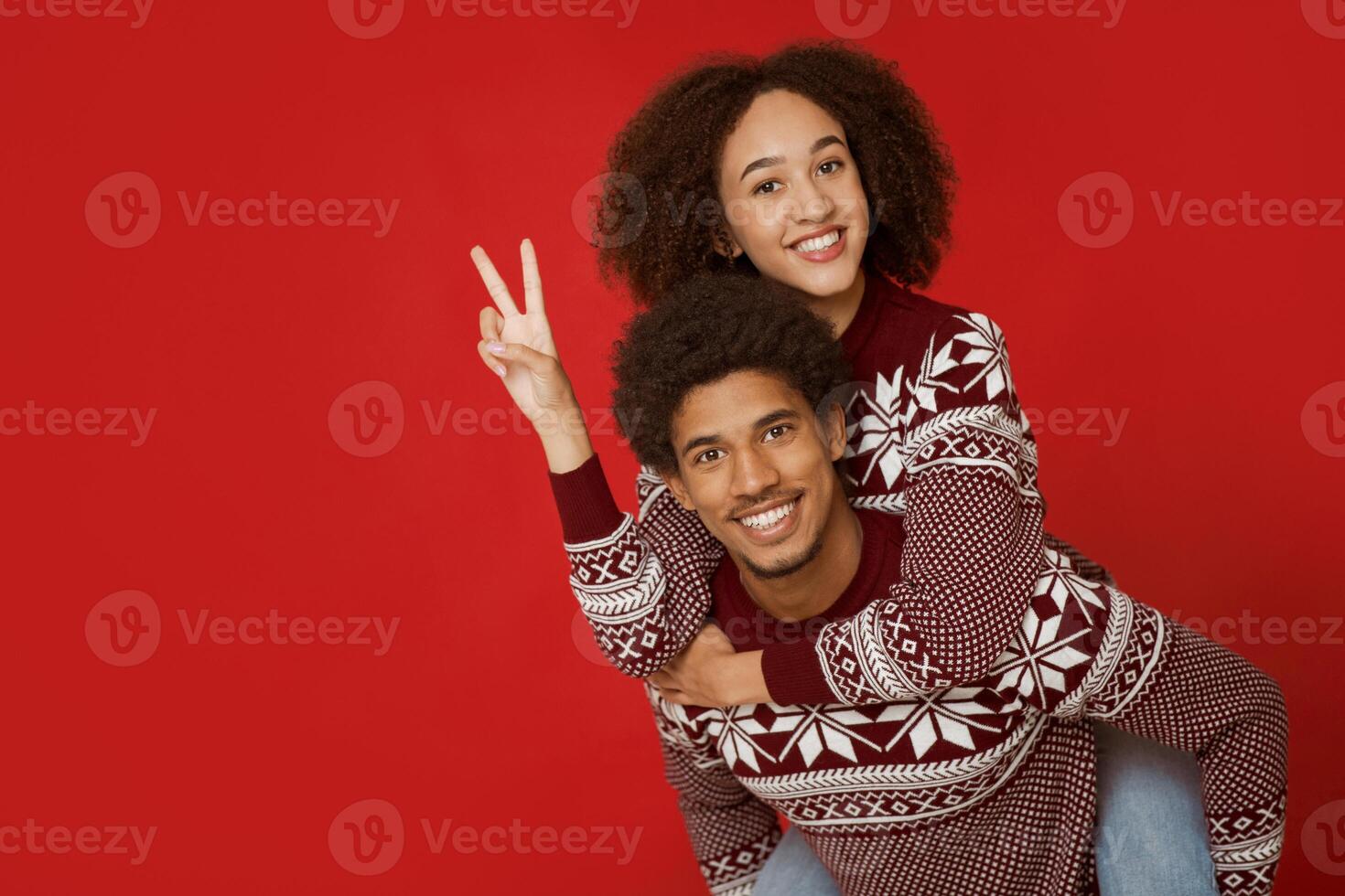 Have fun together during Christmas and New Year holidays. Cheerful young african american male holds female on back, lady shows peas sing with hand and watch at camera, isolated on red background photo