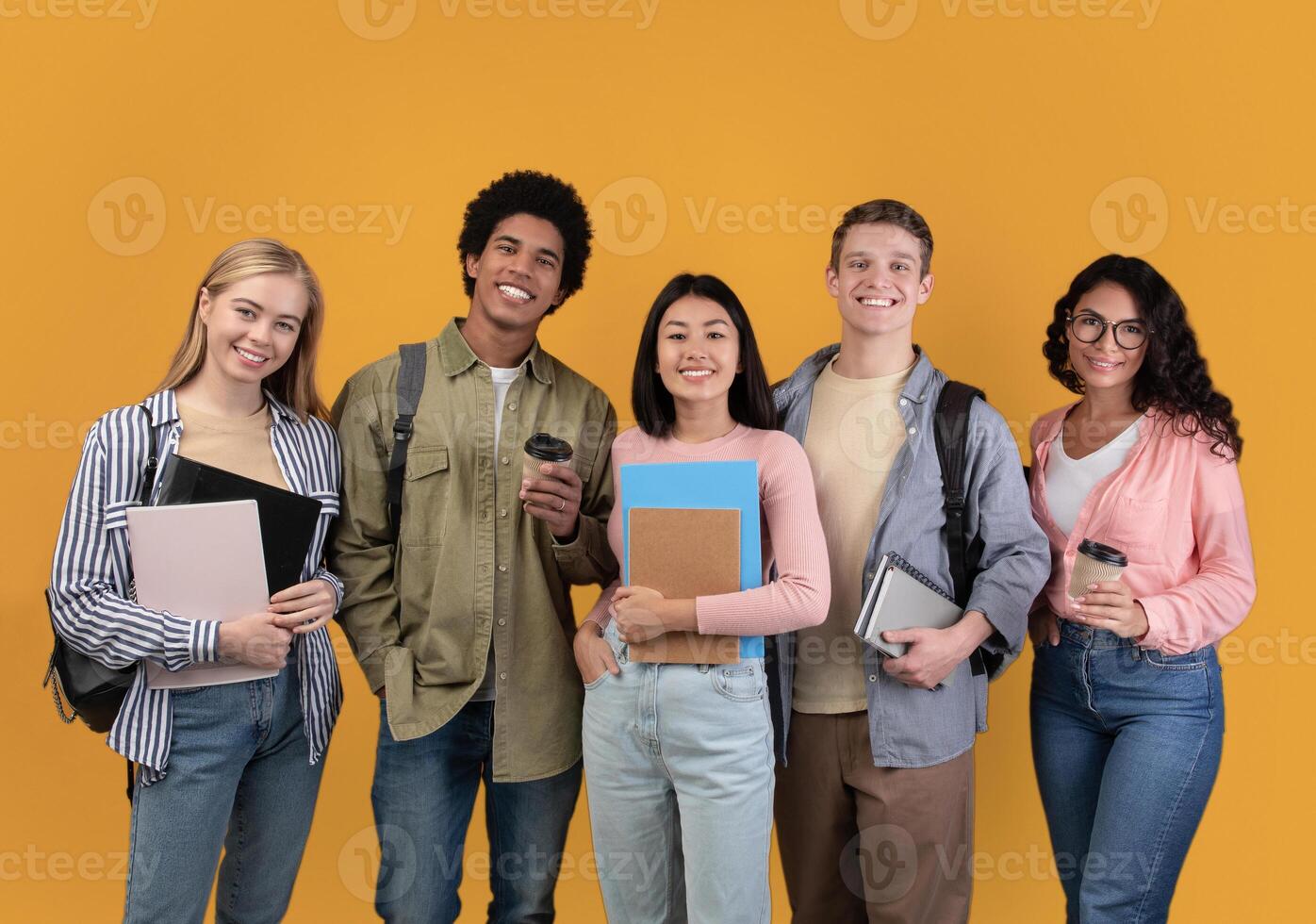 Modern students from different countries and exchange program. Smiling young international people with cups of takeaway coffee, books and backpacks look at camera, isolated on orange background photo