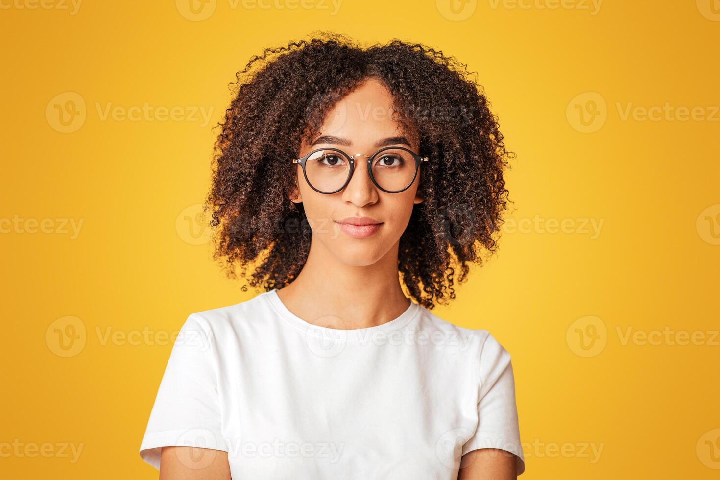 Nerd student ready to study. Smart serious millennial african american lady with curly hair in white t-shirt and glasses looking at camera, isolated on yellow background, studio shot, empty space photo
