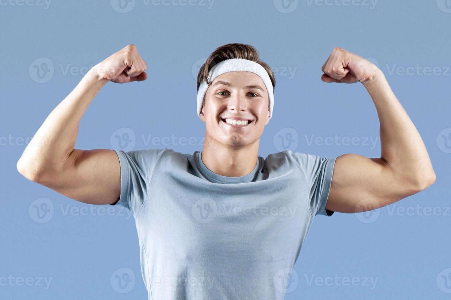 Positive young sportsman sitting on fitness ball and showing his strength on lilac studio background. Attractive millennial guy practicing aerobics or gymnastics, having strong biceps muscles photo