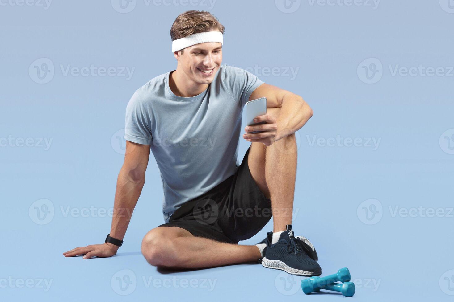 Full length portrait of sporty millennial guy using smartphone while resting after workout on lilac studio background. Fit young sportsman browsing web, checking social media during break photo