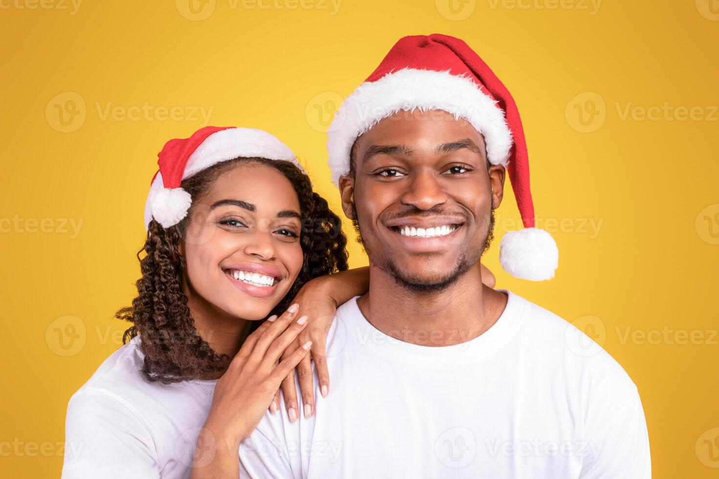 Christmas Portrait Of Joyful Black Couple In Santa Hats Hugging Posing Over Yellow Studio Background, Smiling To Camera. Xmas, New Year And Winter Holidays Celebration. Panorama photo