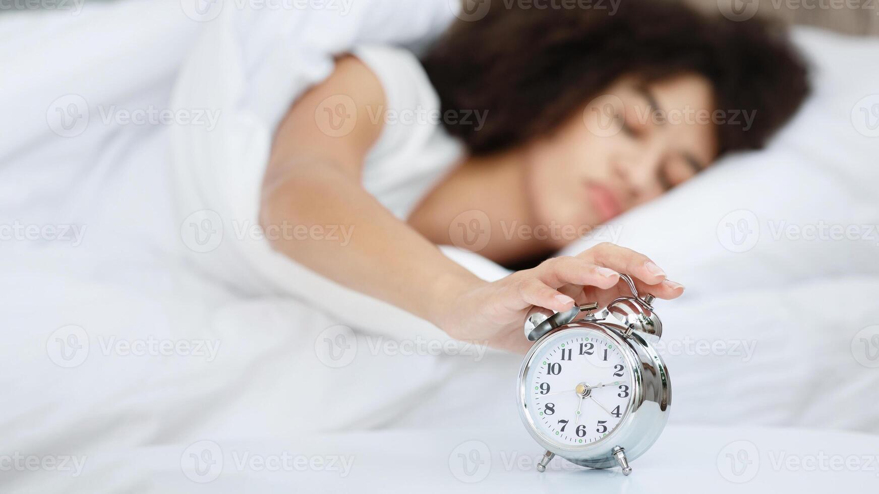 Lady turns off alarm clock, waking up in morning from call. Sleepy millennial african american female lies on bed, turns off alarm and does not want to get up early in modern bedroom interior, blurred photo