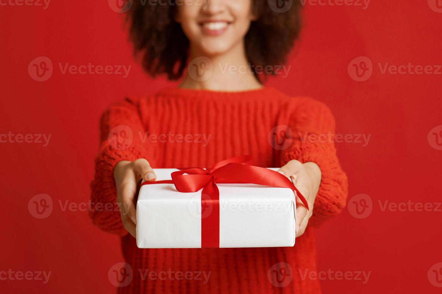 X-mas present, New Year celebration. Cropped studio shot portrait of smiling african american female in sweater gives box with gift, isolated on red background, empty space, close up, panorama photo