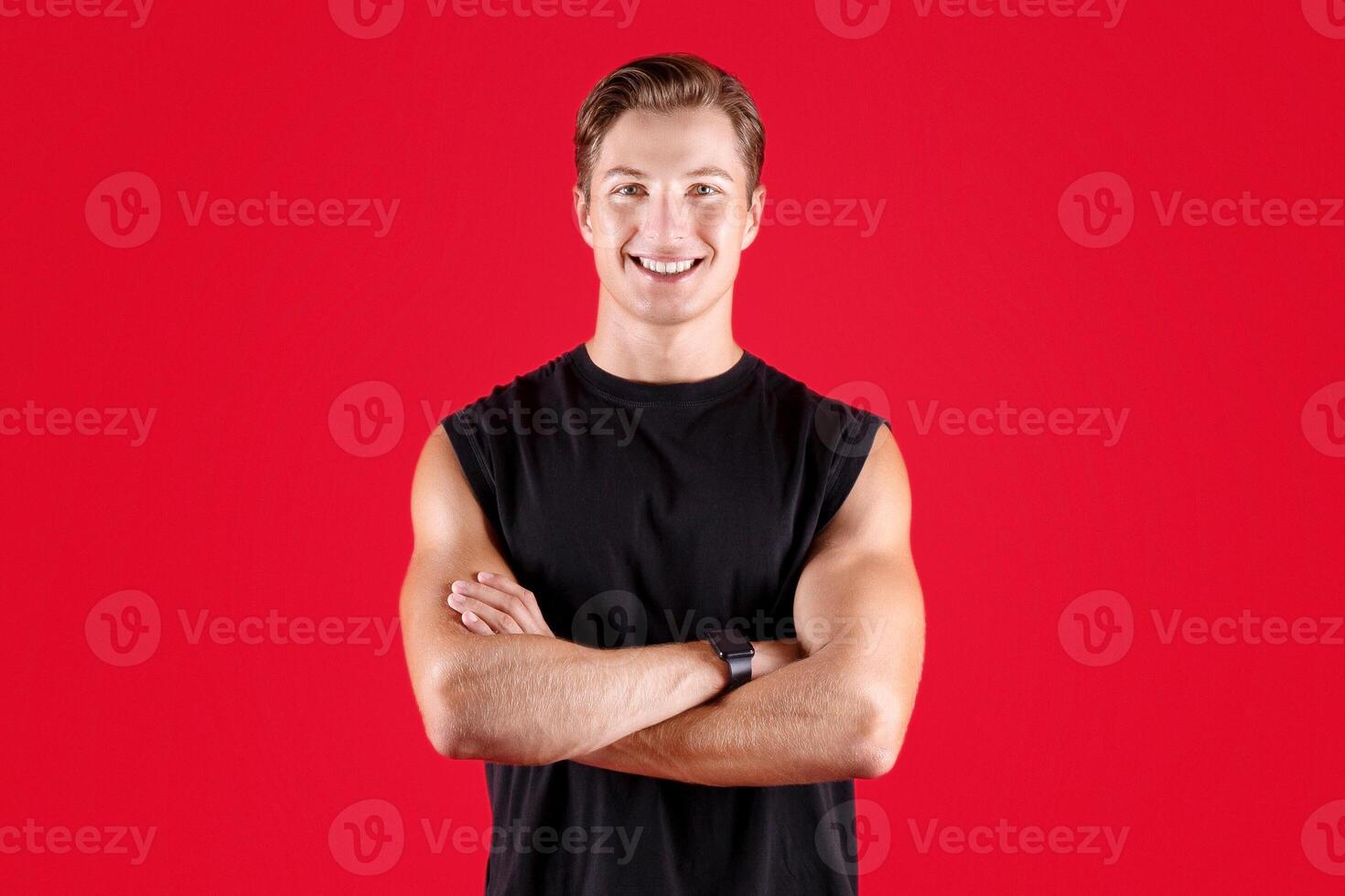 Full length portrait of handsome male fitness model with crossed arms looking at camera on red studio background. Professional young athlete in sportswear posing and smiling photo