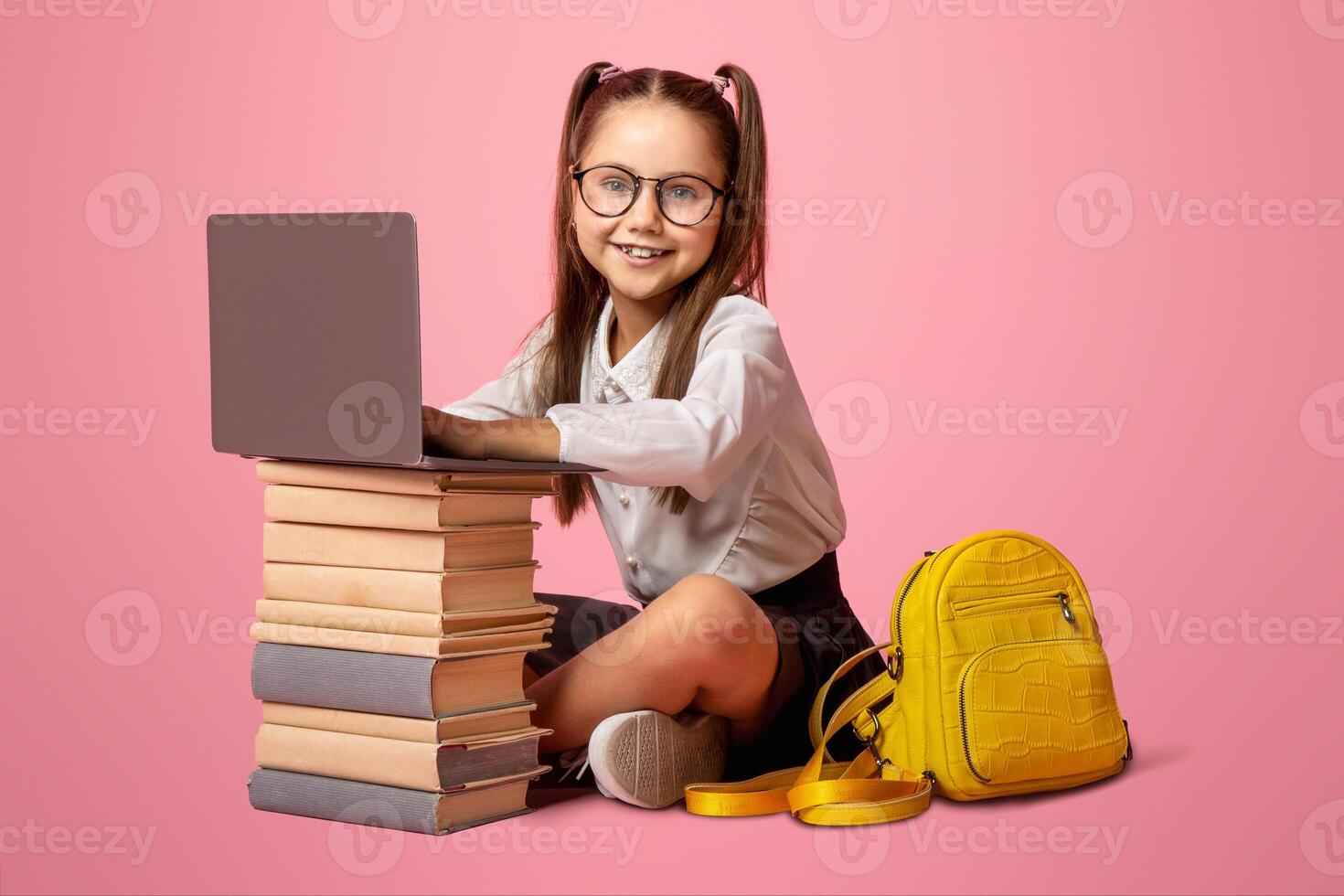 Modern education for children. Smiling girl in glasses with backpack sits near stack of books and laptop, studio shoot photo