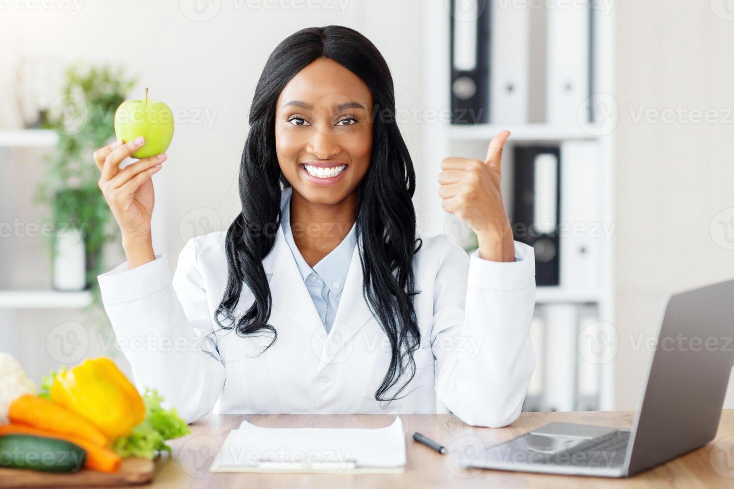Millennial black dietitian holding apple and showing thumb up gesture at desk in medical office. Young female nutritionist recommending weight loss clinic. Healthy eating concept photo