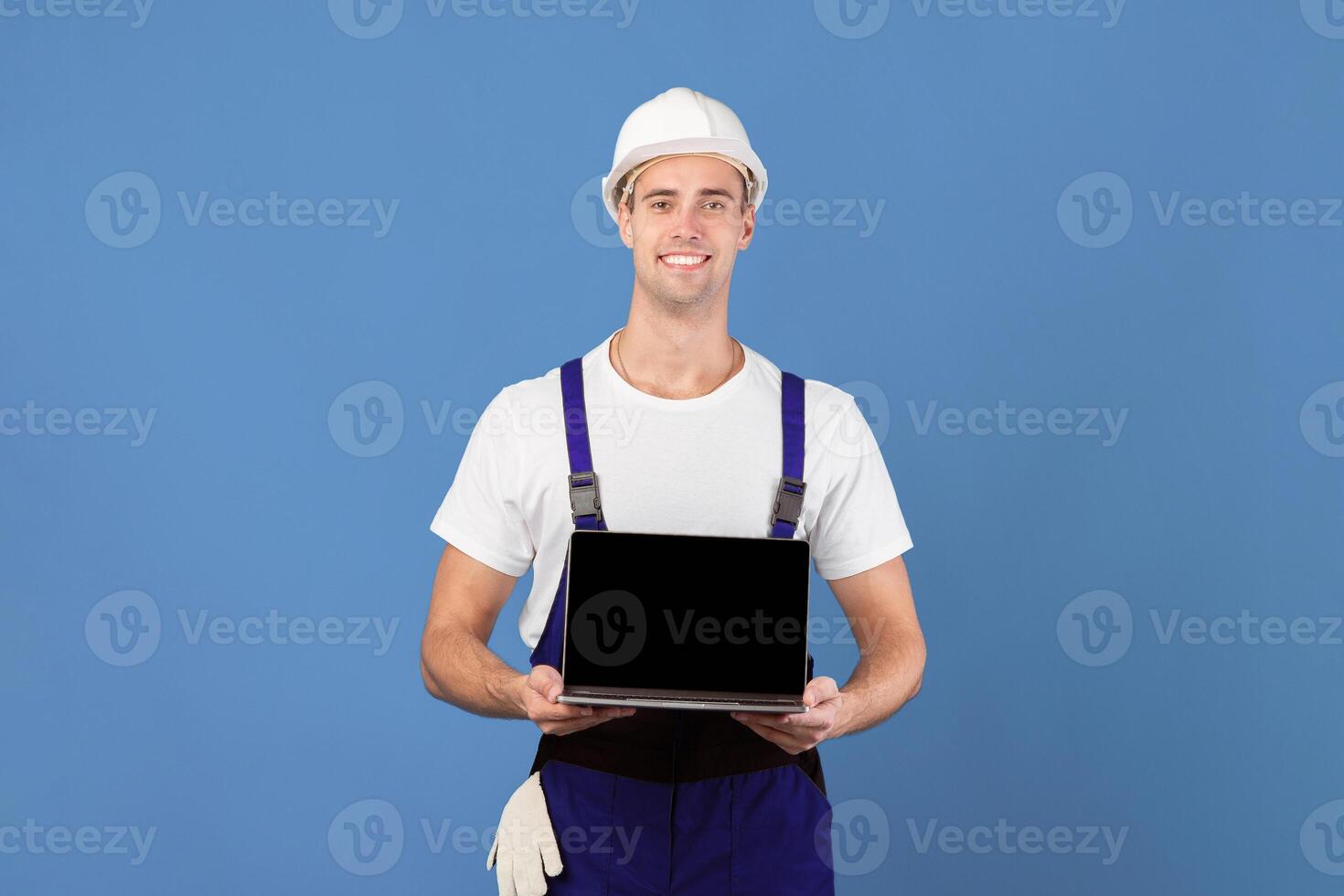 Smiling Foreman Guy In Hard Hat Holding Laptop With Black Screen, Handsome Worker Man Recommending New Website For Online Construction Tools Shopping, Standing On Blue Background, Mockup Image photo