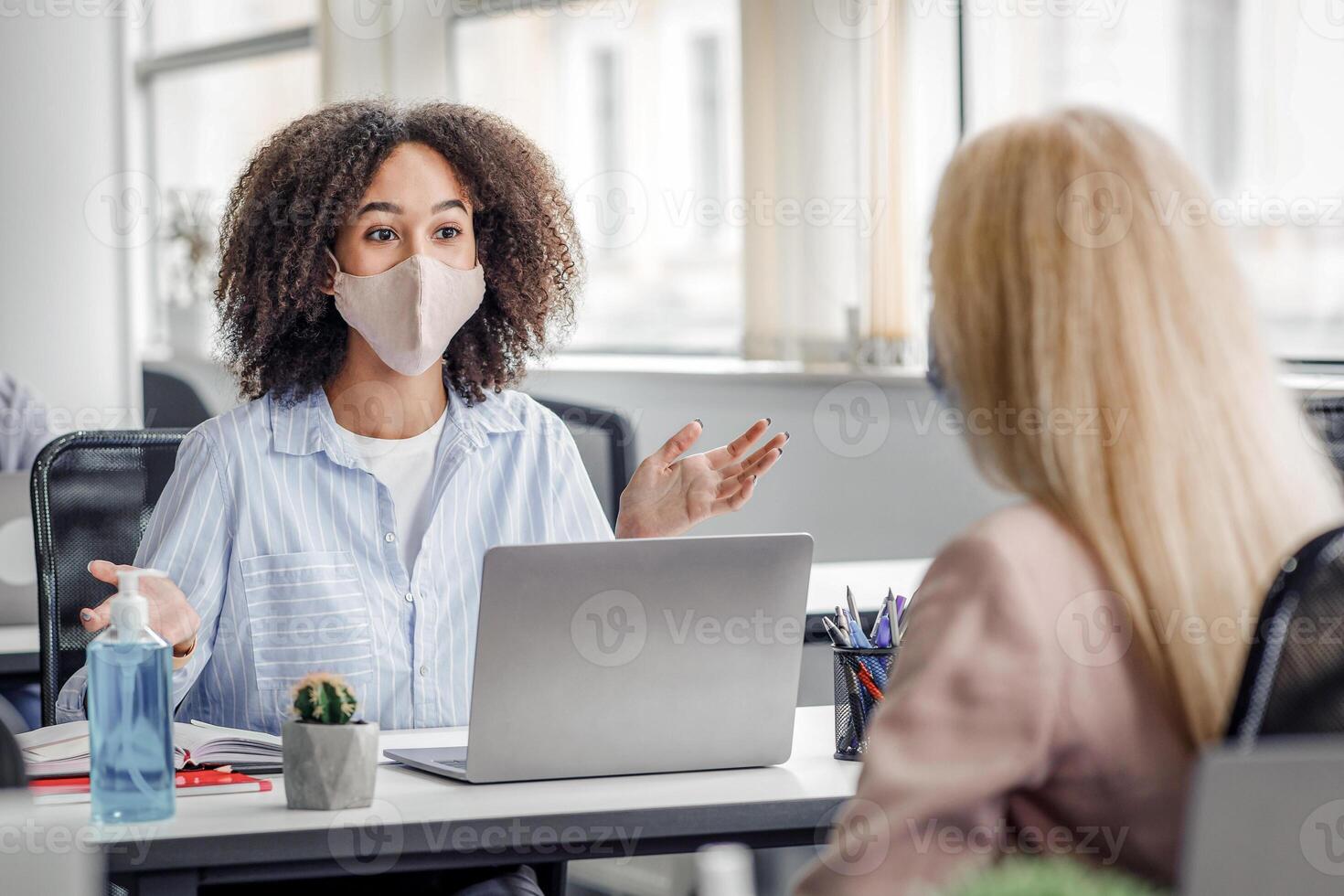 Social distance and communication with client in office during COVID-19 epidemic. African american manager advises woman and gestures at work place with laptop and antiseptic, copy space photo