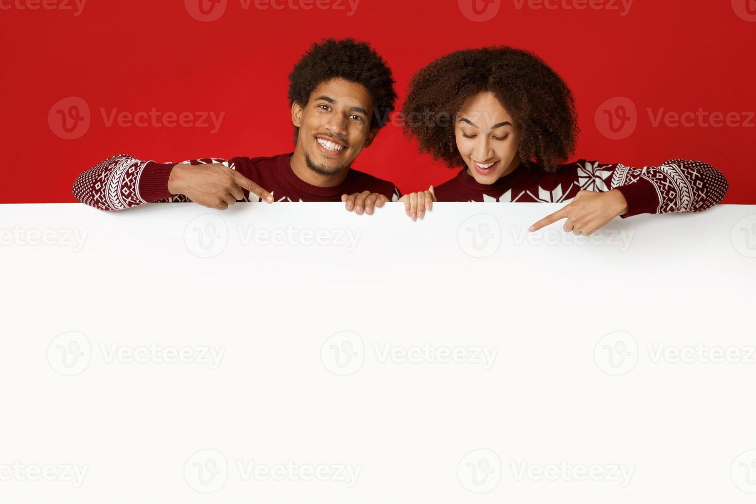 Couple in love advices great proposal for New Year and Christmas. Happy young african american family looking at blank white billboard and pointing with fingers at banner, isolated on red background photo