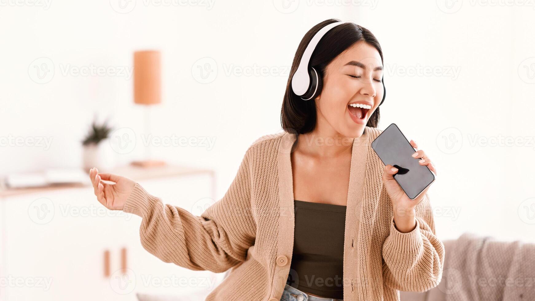 Carefree Singer. Portrait of asian lady singing favorite song and using cell phone as a microphone, dancing and wearing wireless headset. Excited woman having fun at home, enjoying sound, copy space photo