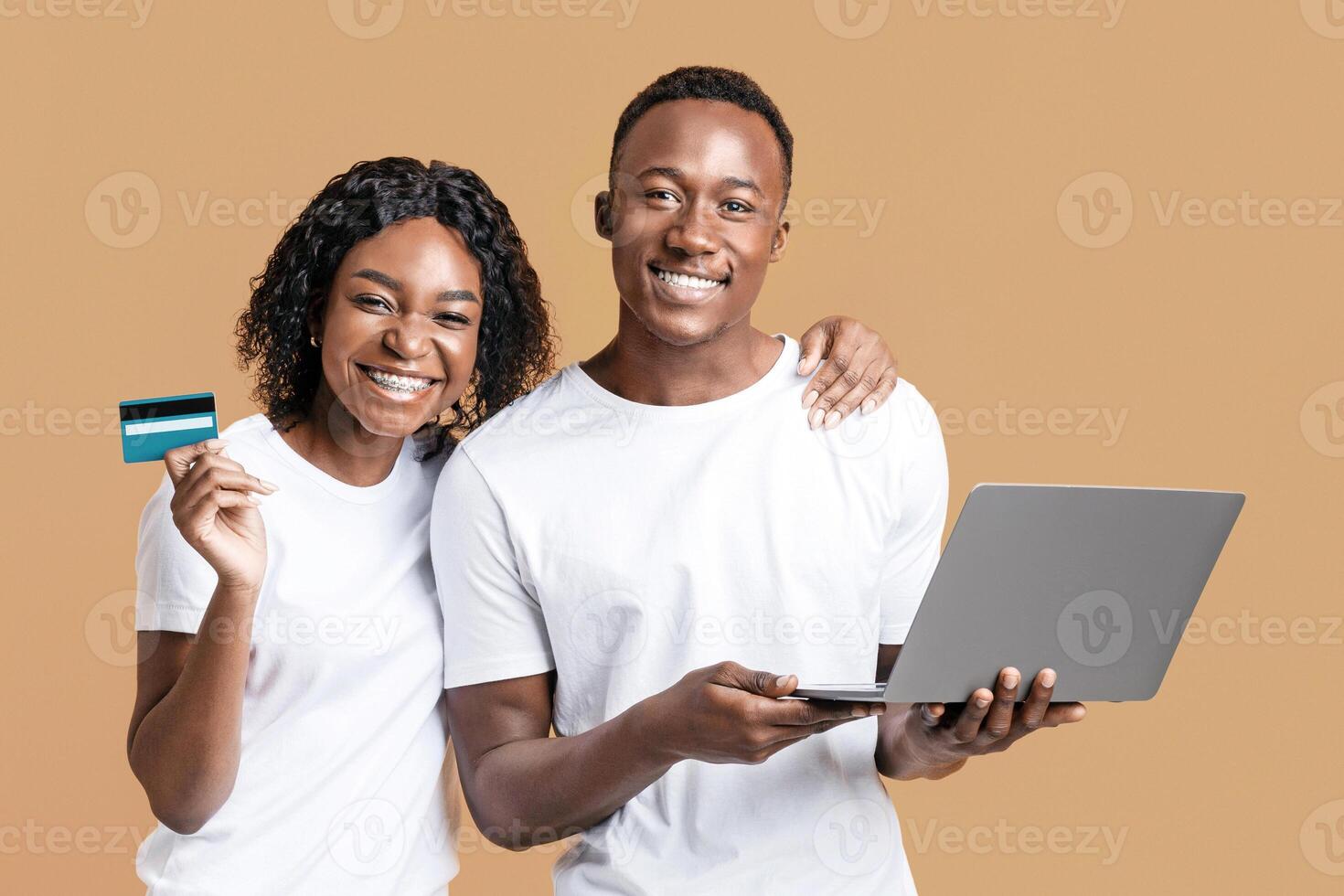 Online banking and e-commerce. Happy african-american man and woman showing credit card and working on laptop on yellow studio background. Black couple making purchases online, using laptop photo