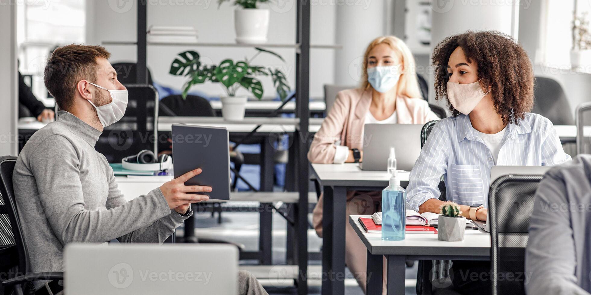 Team of employees working on project and social distancing during outbreak. Millennial male manager in protective mask talk to european and african american women at work desks with laptops photo