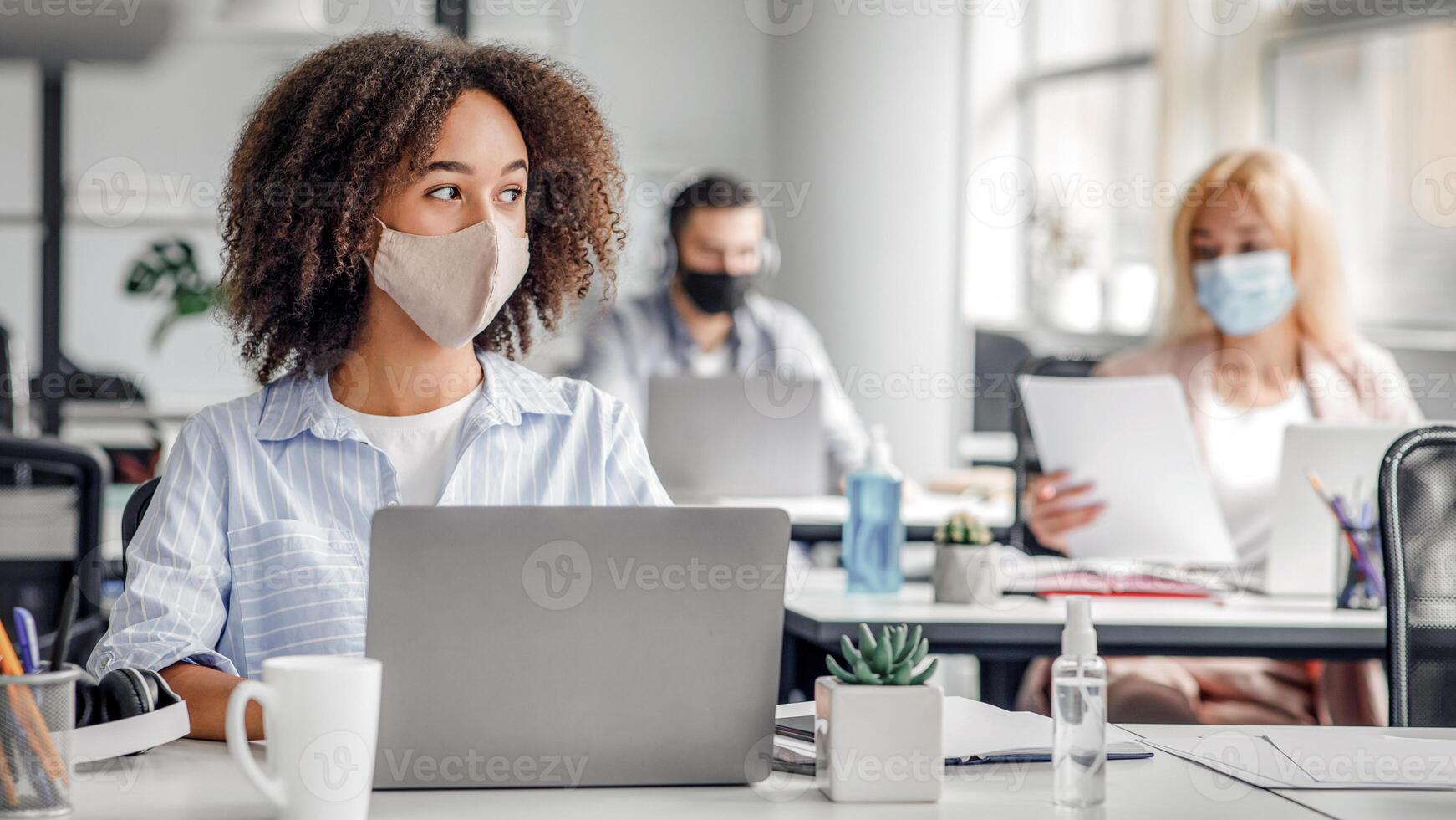 Creating an idea at work brainstorming for project. Serious african american lady in protective mask looks at window working at computer in modern office during outbreak of coronavirus, copy space photo