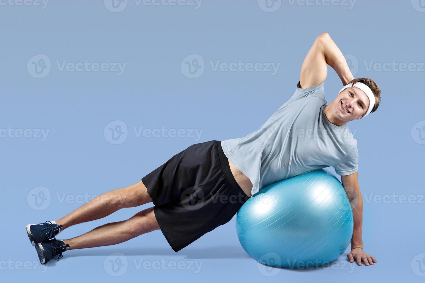 Athletic millennial guy doing side plank with fitness ball over violet studio background, space for text. Full length portrait of happy young man keeping in good shape, doing aerobic workout photo