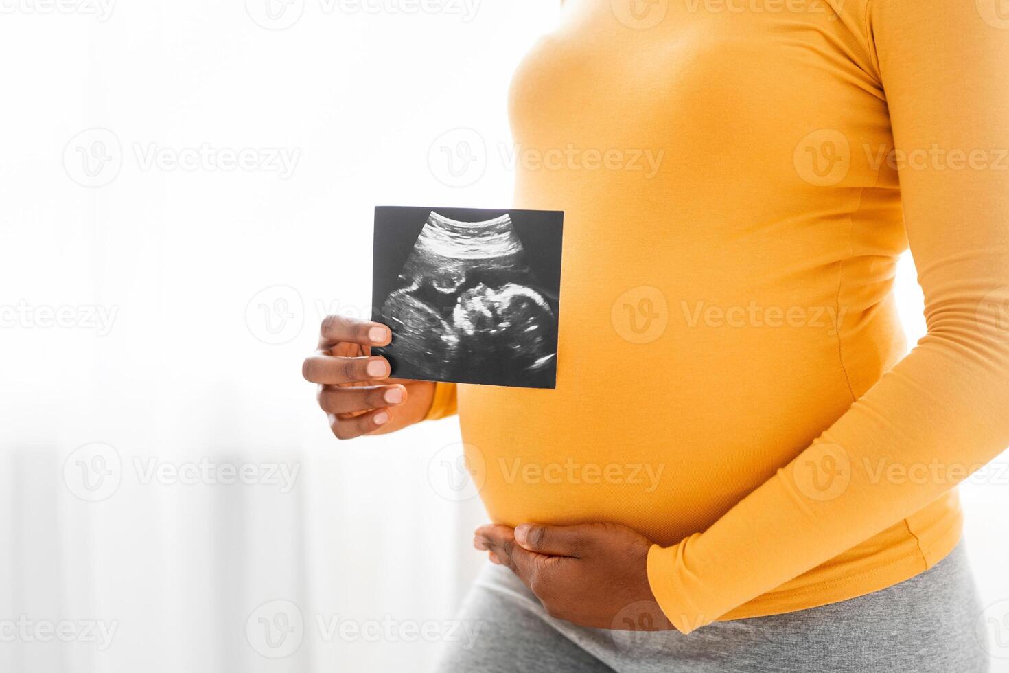 Pregnant black woman showing her baby sonography, enjoying future motherhood with first photo of her unborn child, free space. Unrecognizable african mother standing by window at home