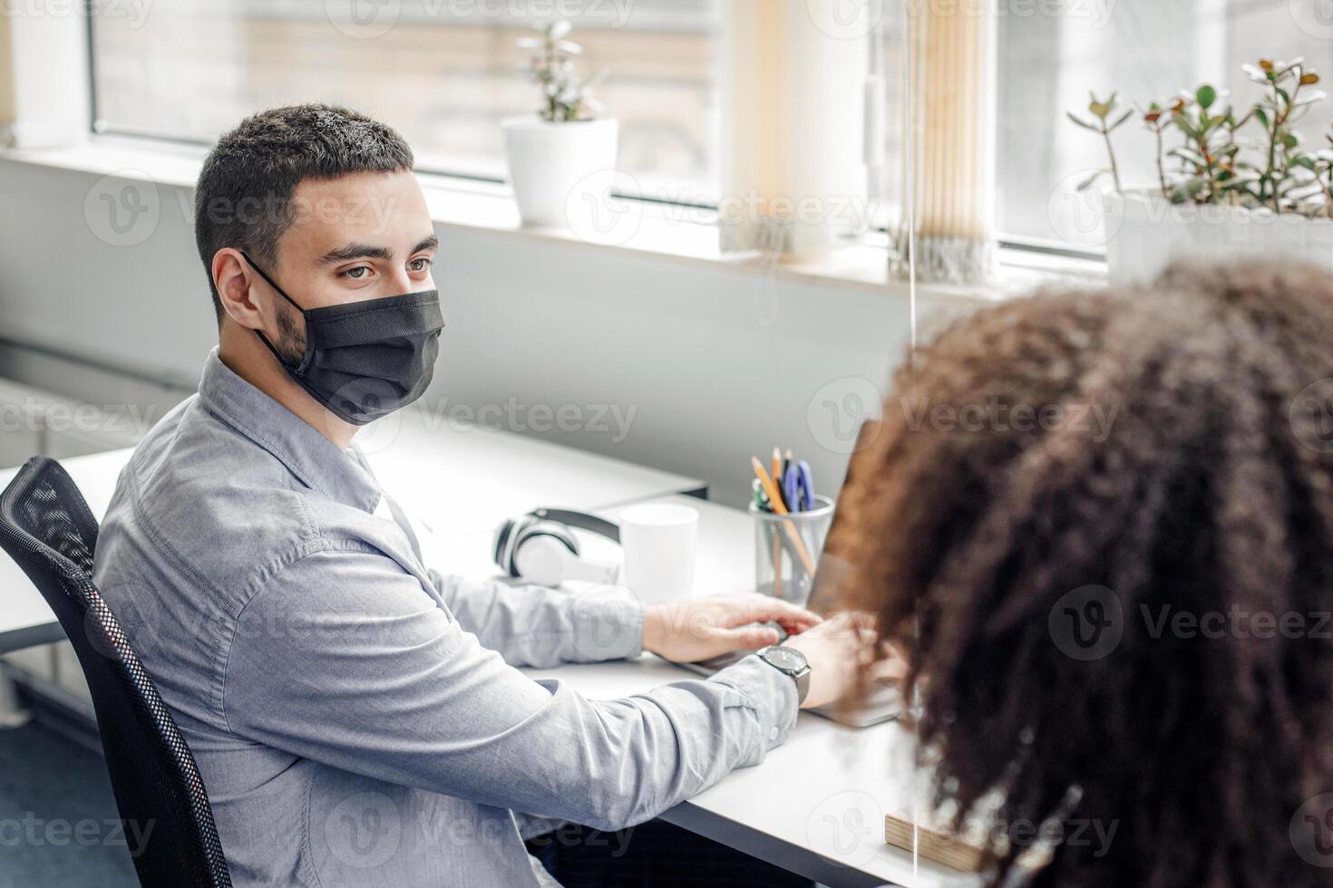 Social distance and communication at work with colleague during epidemic. Guy in protective mask and african american lady talking at workplace with laptops in office through protective glass photo