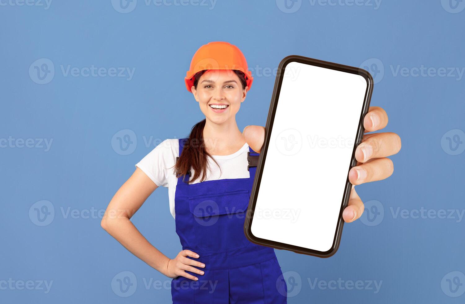 Smiling young woman in coveralls and protective hard hat holding paper v-sign check mark symbol while posing over blue studio background, positive handywoman ready for repairment works, copy space photo