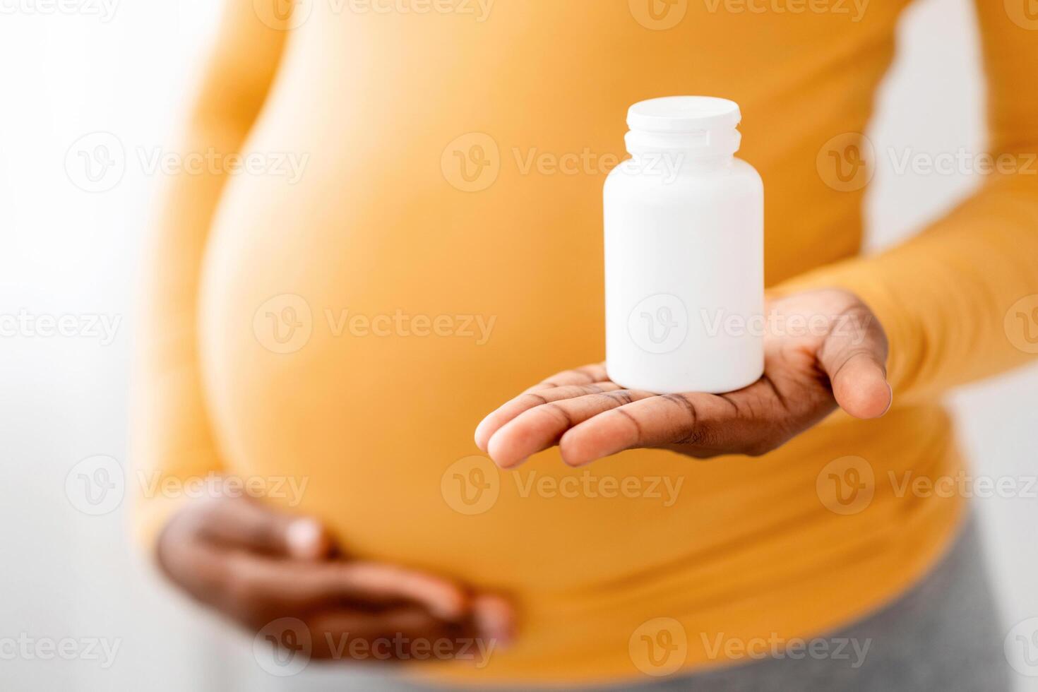 Closeup of black pregnant woman showing jar with pills, supplements or vitamins and touching her big tummy. Unrecognizable african american expecting lady recommending supplements for future mothers photo