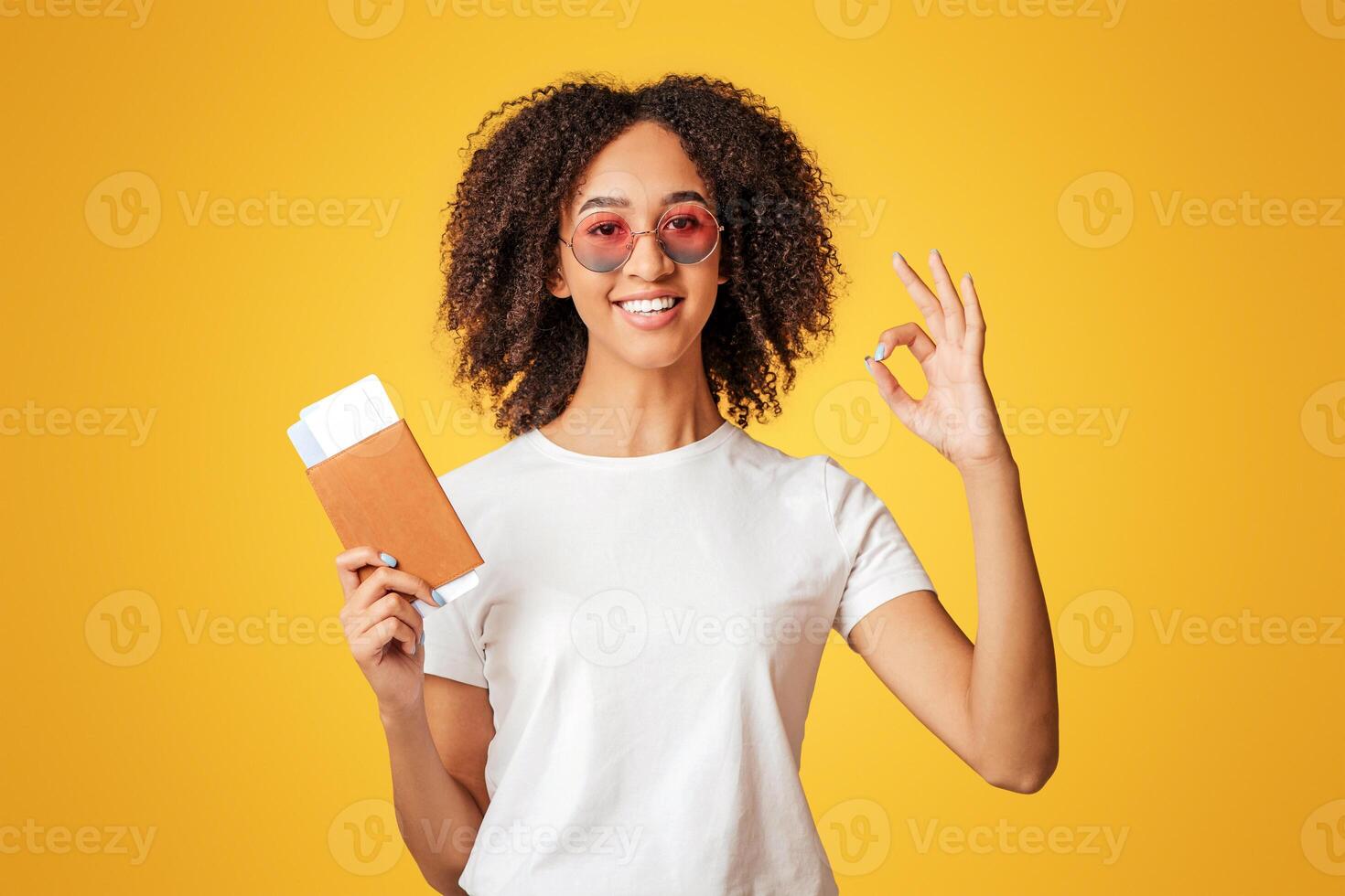 Travel, air journey and tourism after covid-19 lockdown. Happy millennial african american female with curly hair in white t-shirt with glasses holds passport with tickets and shows signs okay photo