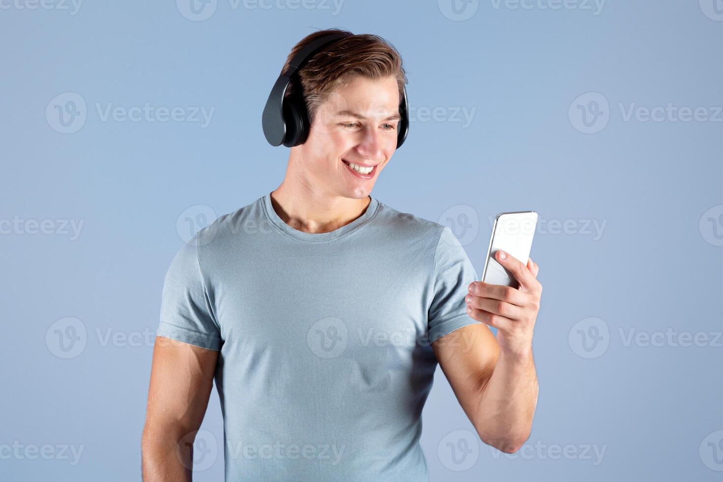 Young sporty guy with smartphone listening to music in headset on violet studio background. Happy millennial sportsman choosing playlist for his training on mobile device photo