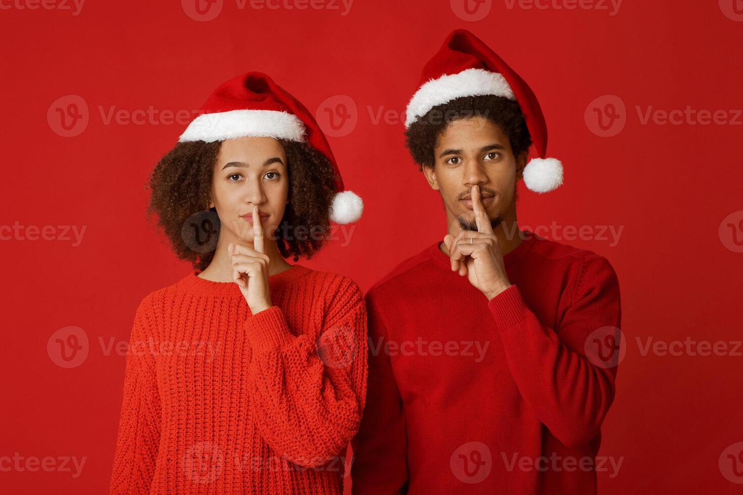 Secret Santa and wish on New Year and Christmas. African american young couple in sweaters and Xmas hats makes sign of silence and watch at camera, isolated on red background, panorama, studio shot photo