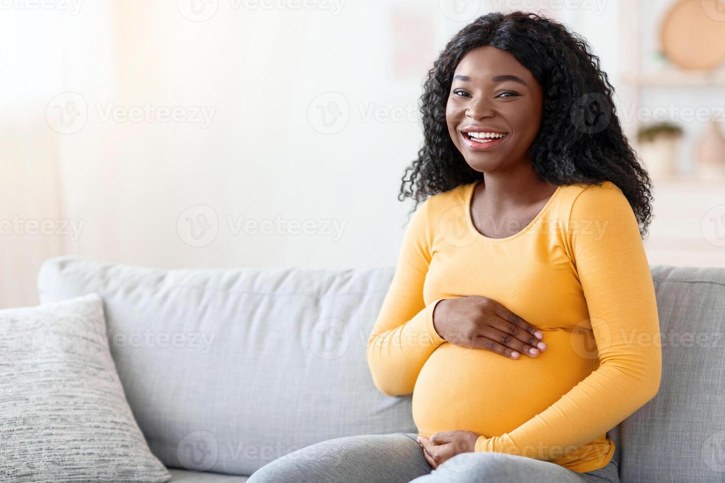 Joyful black pregnant woman hugging her big tummy, communicating with her baby, sitting on sofa at living room. Happy african american expecting lady spending time at home, looking at copy space photo