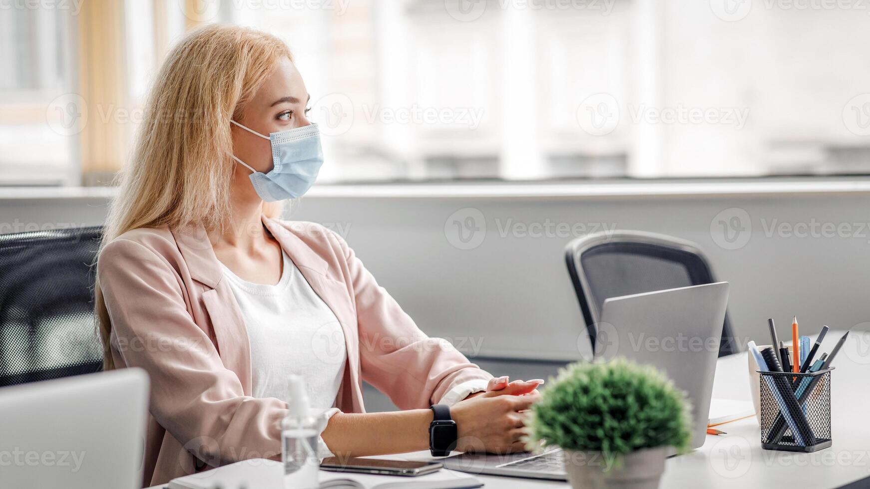 Business must be continuous, healthcare concept. Blond woman in protective mask at workplace with laptop, antiseptic and smartphone looks out the window in office interior during pandemic, free space photo