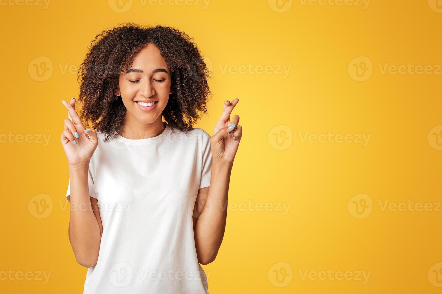 Make wish, luck and dream come true. Happy young african american lady in white t-shirt with curly hair, with closed eyes crossing fingers, isolated on yellow background, studio shot, empty space photo