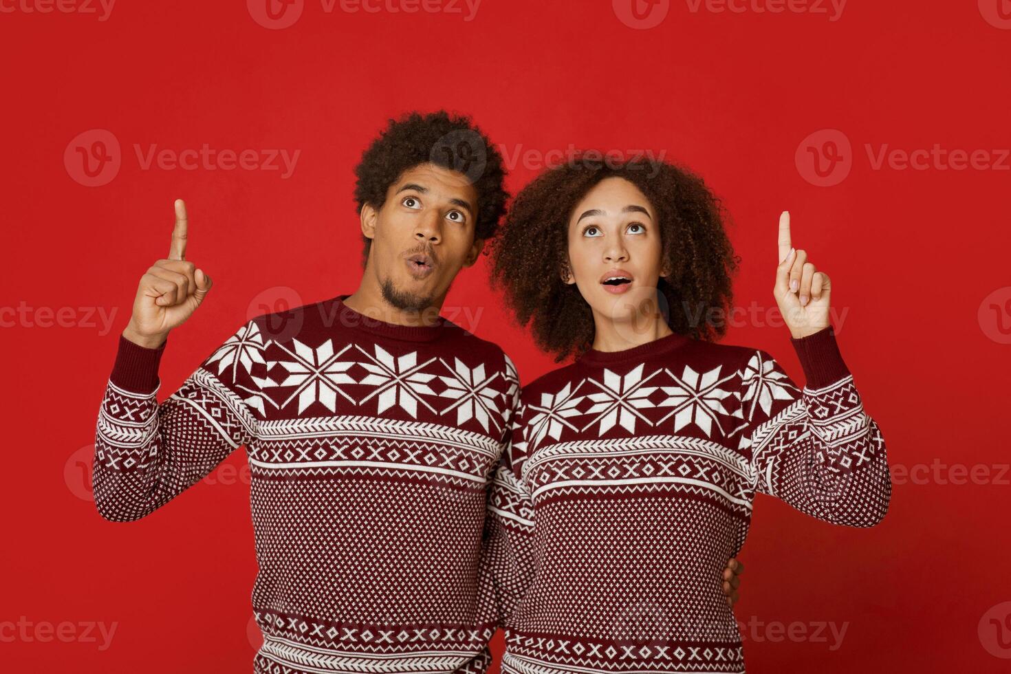 Amazing offer for gifts for New Year and Christmas. Surprised african american couple in sweaters pointing up and looking at free space, woman with open mouth isolated on red background, studio shot photo