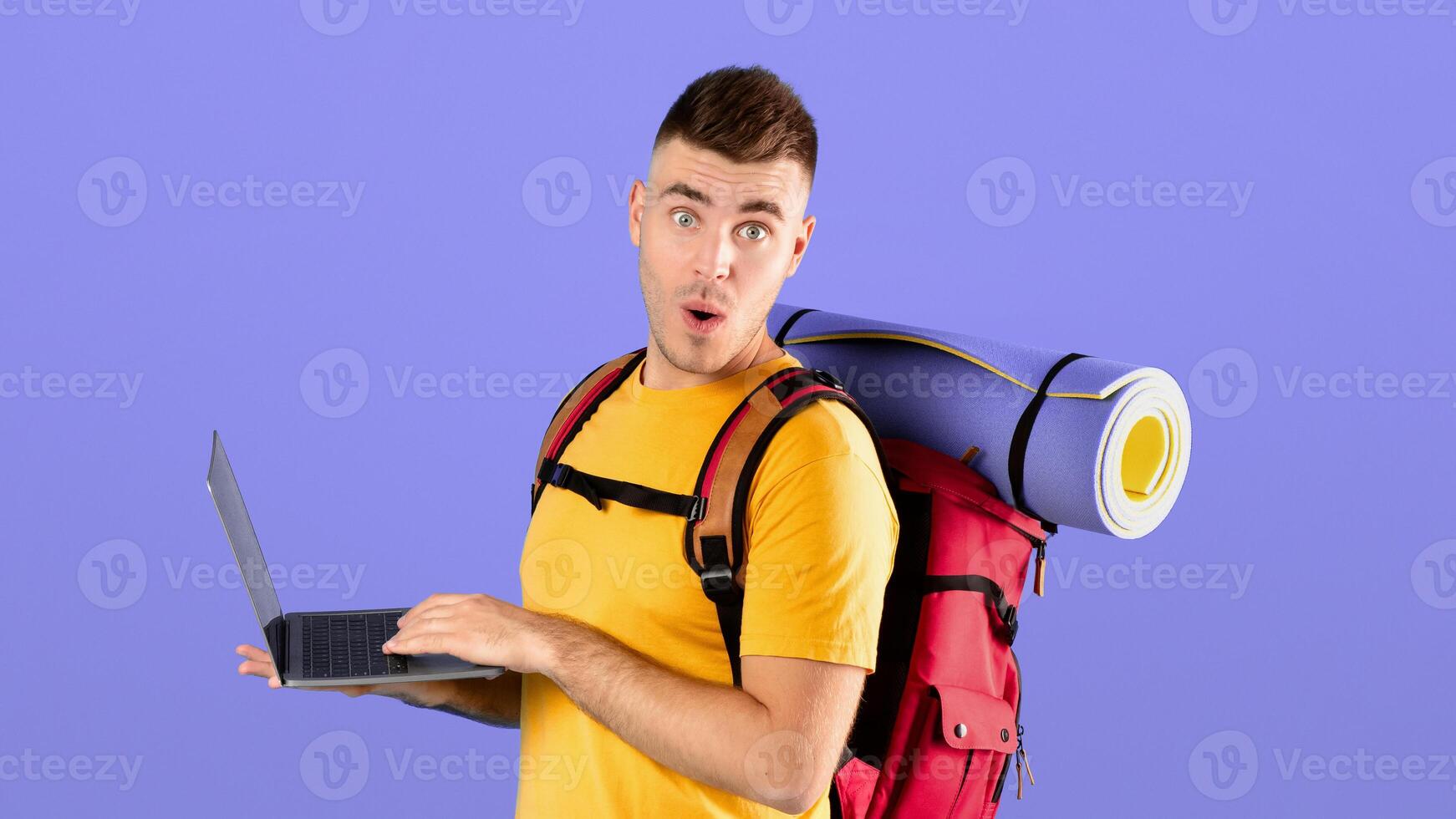 Booking tour online. Young guy with camping gear and laptop surprised over big vacation discount in online travel agency, blue studio background. Shocked hipster purchasing weekend journey on web photo