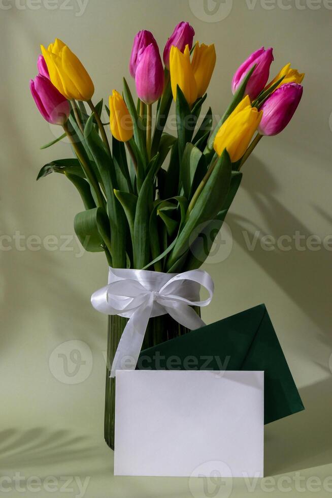 Empty white envelope mock up template Multicolored fresh tulips in vase in green background with trendy deep shadows. Spring flowers in yellow and violet colors. Postcard wallpaper design Copy space photo