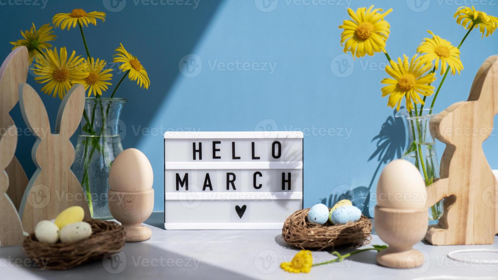 Lightbox with message HELLO MARCH on festive blue background and eco-friendly zero waste natural decoration. Yellow daisies flowers. Bunny egg. Holiday greeting card photo
