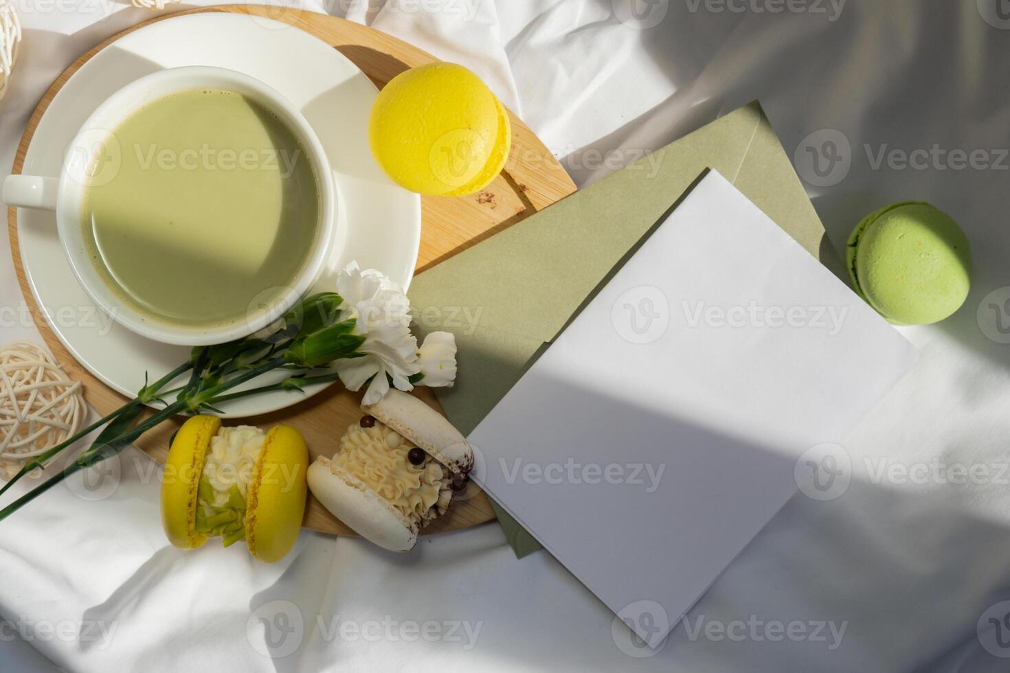 Copy space letter mock up empty envelope Nutrient-rich matcha drink with trendy macaron dessert on wooden tray in bed with fresh flowers. Morning beautiful breakfast present gift idea. Vibrant creative colors. Natural energy givers tea photo