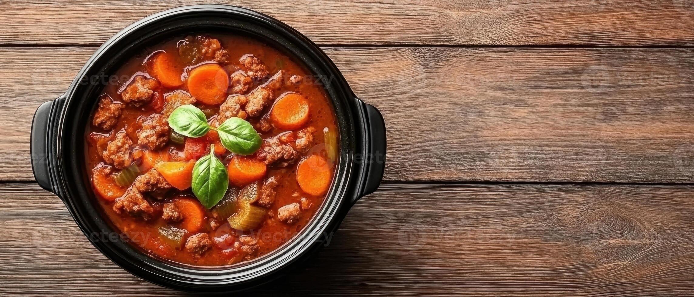Hearty meat and vegetable stew in a black pot. Fresh basil leaves garnish the dish on a brown wooden table background photo
