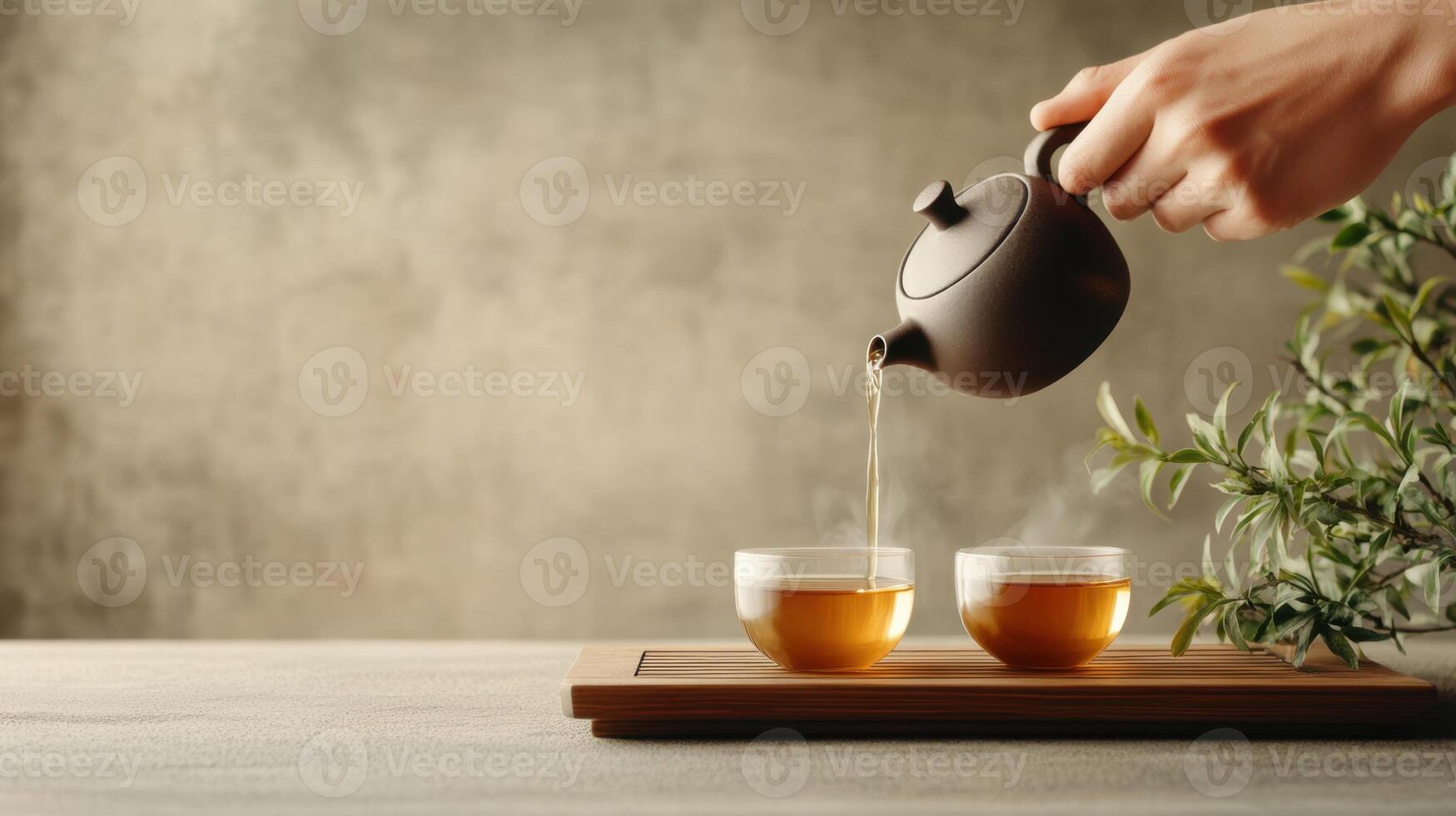 Hand pouring hot tea from brown teapot into clear glass cups on a wooden tray with plant against grey textured background photo