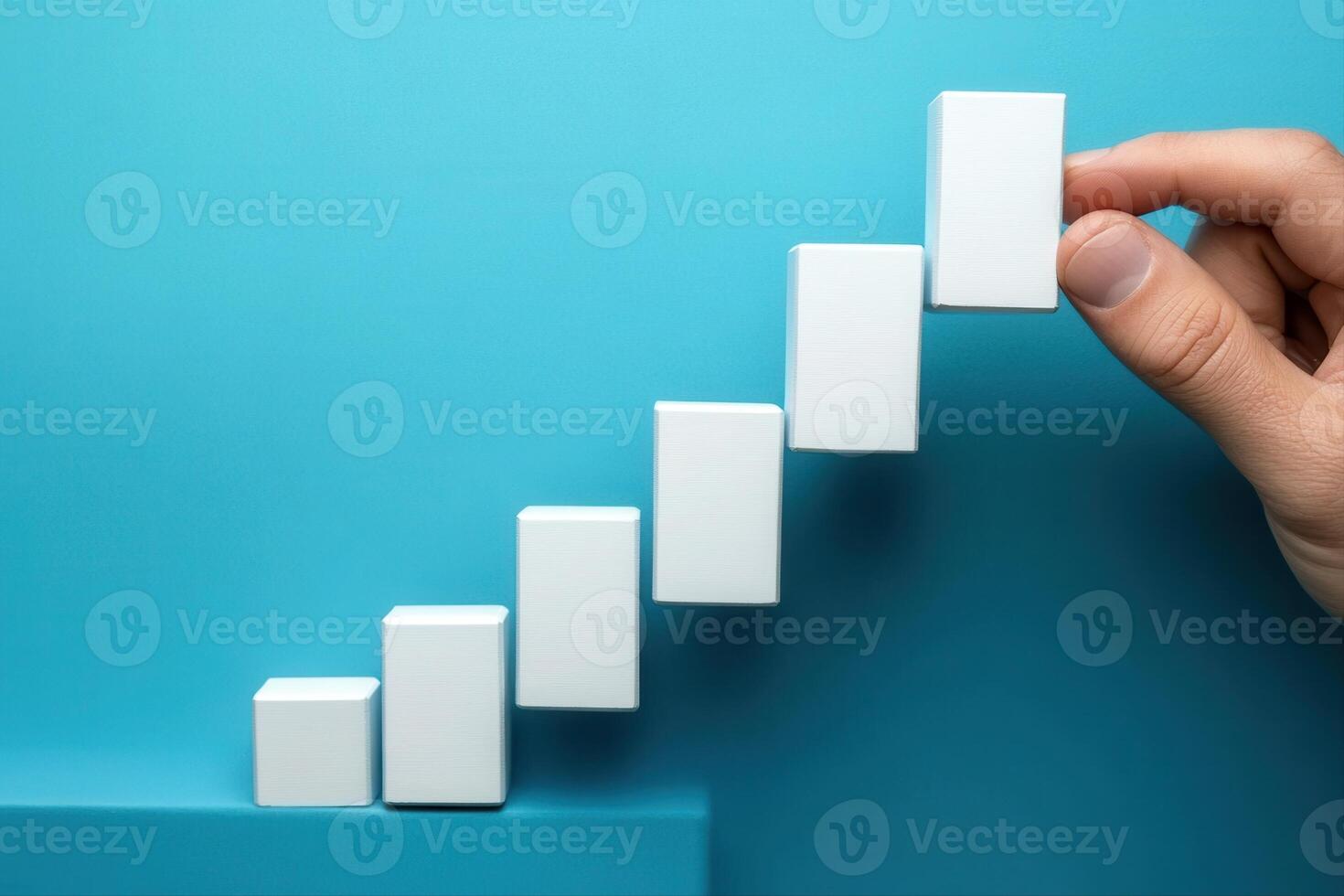Hand placing a white block on a staircase of blocks against a blue background, symbolizing growth photo