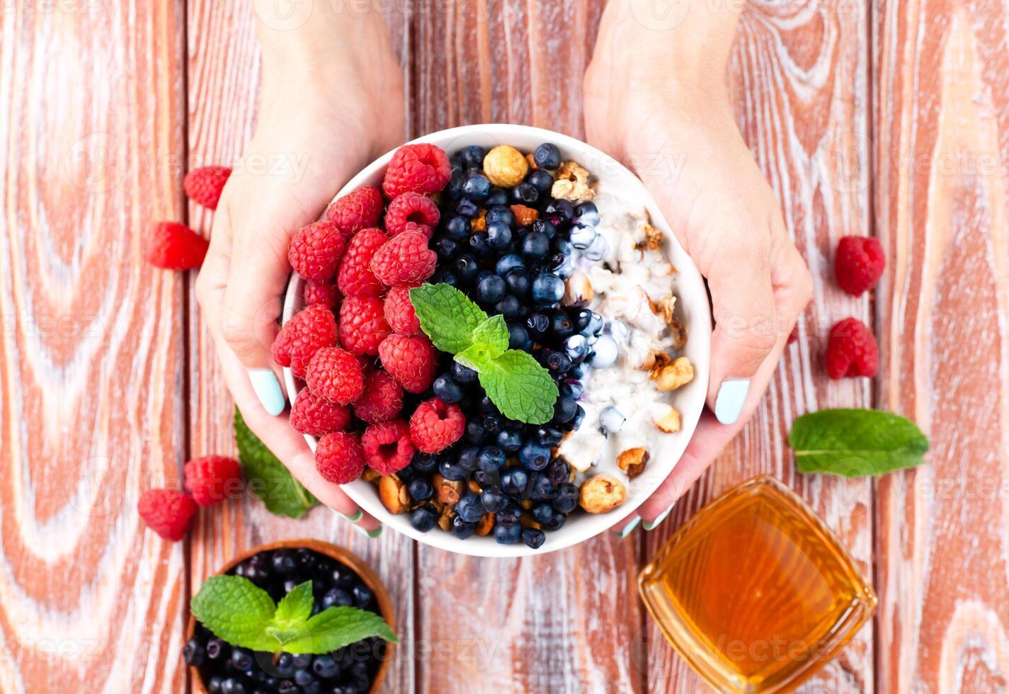 The girl's hands hold a bowl of muesli with ripe berries, yogurt and honey on the wooden table. Healthy breakfast. Top view. photo