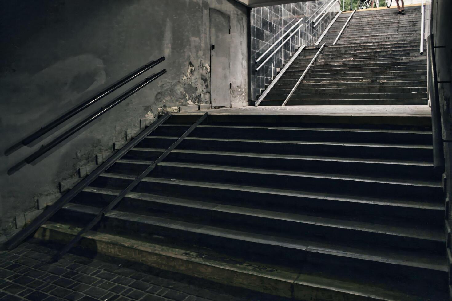 A dimly lit underground passage with two sets of stairs leading upward. The worn walls, peeling paint, and metal handrails create an eerie, abandoned atmosphere, with a bicycle faintly visible at exit photo