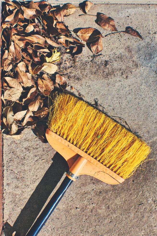 Yellow broom resting on a concrete surface with scattered dry leaves. Ideal for autumn, cleaning, or outdoor themes, capturing a rustic, seasonal moment. photo