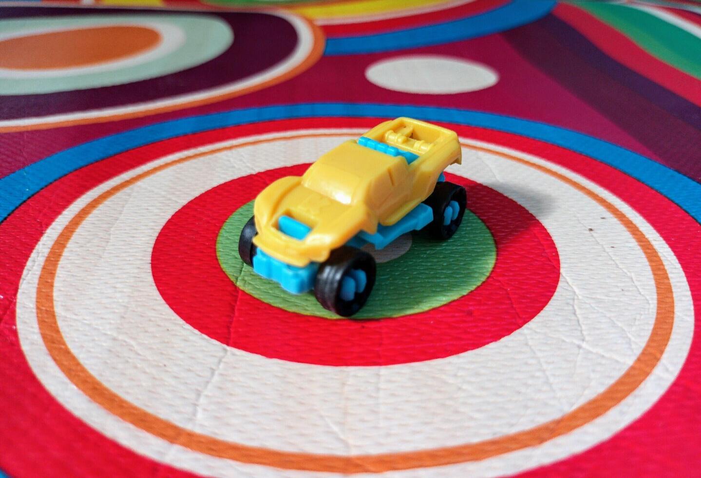 A Small Yellow and Blue Toy Car on a Colorful Circular Patterned Mat. photo