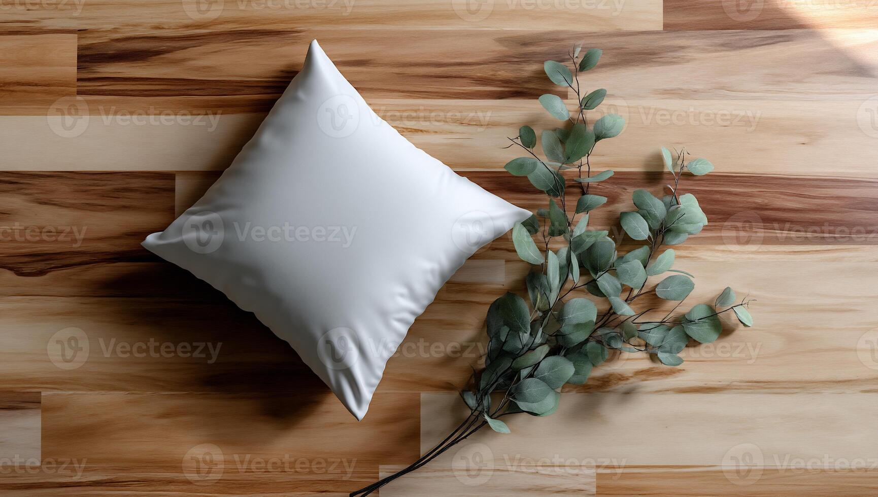 A plain white square pillow rests on a light brown wooden floor, beside a eucalyptus branch photo