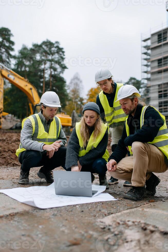 Construction team reviewing blueprints on-site. Several construction workers in safety vests and hard hats, huddled around a laptop and blueprints, focused on project plans on a construction site photo