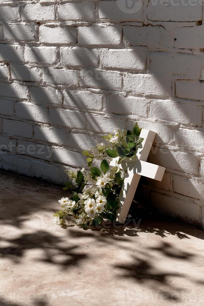 Simple white cross adorned with flowing white flowers and greenery, against a sunlit white brick wall photo