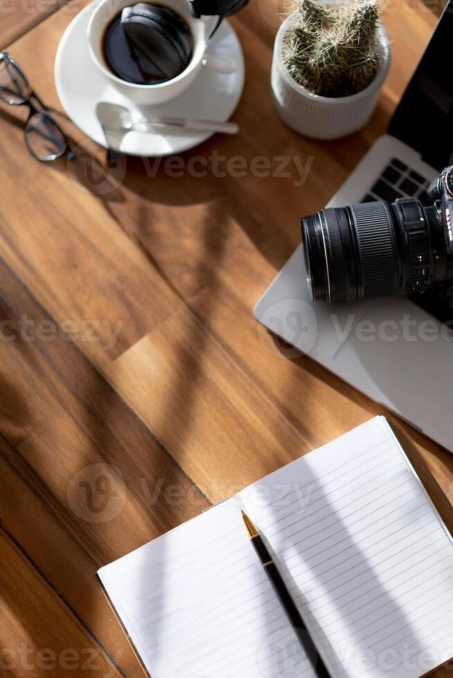 Workspace with coffee, camera, laptop, and notebook. Sunlight casts shadows on wood table photo