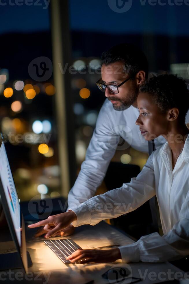 Two colleagues collaborate late at night on a computer, reviewing data or a presentation. City lights visible outside the window photo