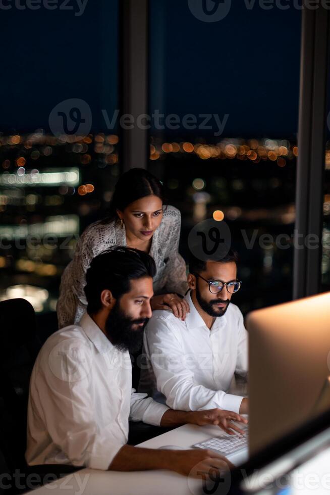 Night work team collaborates on a computer. Focused professionals work late into the night, overlooking city lights photo